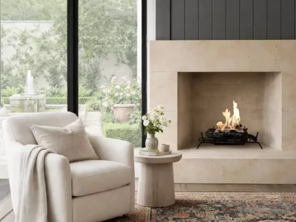 Cozy modern living room with a light upholstered armchair beside a lit stone fireplace, large black-framed windows, and a patterned area rug.